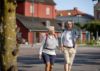 Ett äldre par håller varandra i handen på ett torg med ett rött hus i bakgrunden.