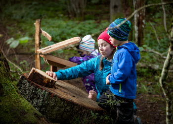 Mamma med två barn utforskar trädets ringar ute i skogen. De har alla mössor och friluftskläder på sig.