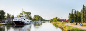 En stor vit passagerarbåt ligger vid Töreboda gästhamns brygg. Göta kanal syns och en promenadväg längs kanalen.