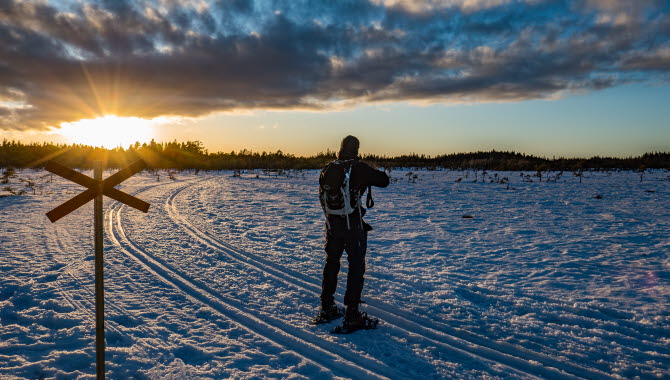 En man i snöskor står i solnedgången och blickar ut över ett snöklätt Blängsmossen.