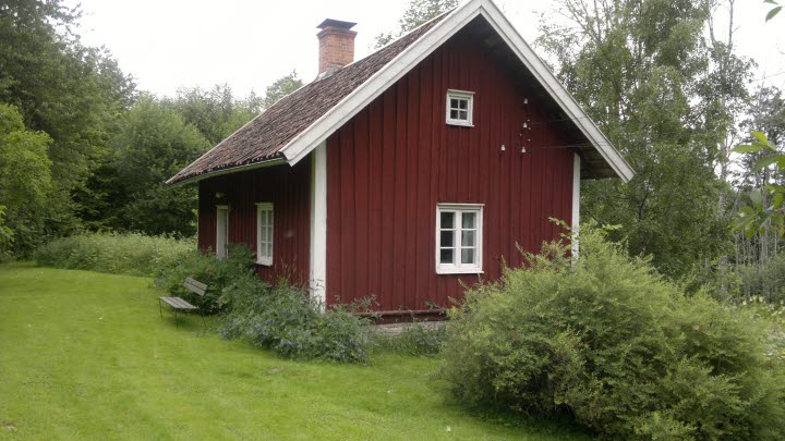 En röd stuga med vita knutar på en grönskande tomt.