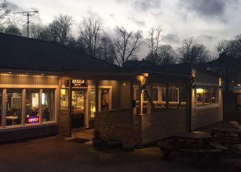 En bild på en resturang med ett trädäck för uteservering på framsidan. Här lyser många lampor, ljusslingor och neonskyltar i skymningen