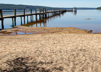 En bild på en sandstrand intill en stor blå sjö- Himlen är klarblå och molnfri. En stor, rejäl brygga går rakt ut i vattnet. Längst ut på bryggan ligger en båt.