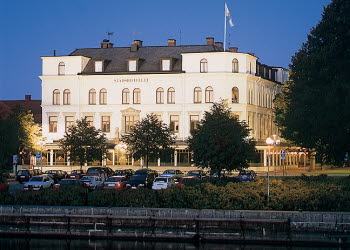 A large old white building with black roof right next to a river. It is evening and the lighting illuminates the building in a beautiful way.