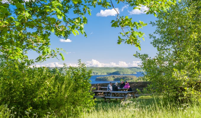 En bild på gröna löv i förgrunden, följt av ett sällskap som fikar vid ett bord. I bakgrunden är en otrolig utsikt mot en sjö.