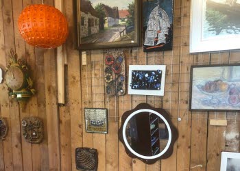 En trävägg fylld med gamla tavlor, lampor, klockor och speglar. Framför väggen står fyra mindre sidbord.