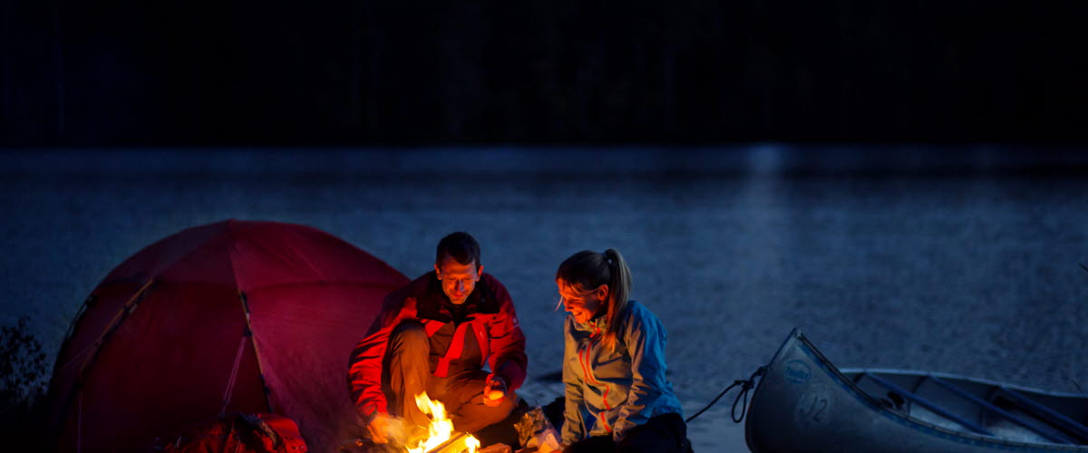 Ett par sitter vid utanför sitt tält vid en eld på kvällen. Bakom dem ser man en sjö och en skog. En kanot ligger vid sjökanten.