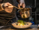 Plating a dish, pan and sauce spoon is held by a pair of hands.