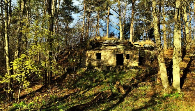 En grotta ute i skogen, med två små öppningar som fönster och en större som dörr. Solen lyser och träden skapar skuggor i marken.