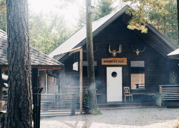 En bild på tre svartmålade små stugor som stor omringade av frodig skog.