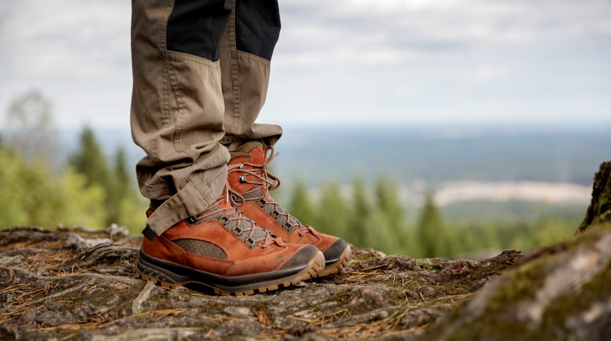 The picture is showing someones hiking shoes and hiking trousers. The person is standing on a height with a view  in the background.
