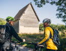 Two people are standing next to their bicycles. In the background, Södra Råda timber church is visible