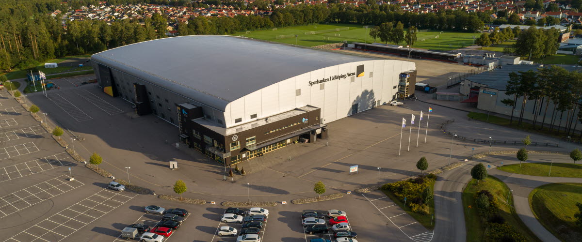 Sparbanken Lidköping Arena som är en grå stor arena. En stor parkering framför arenan och bakom är det fotbollsplaner. 