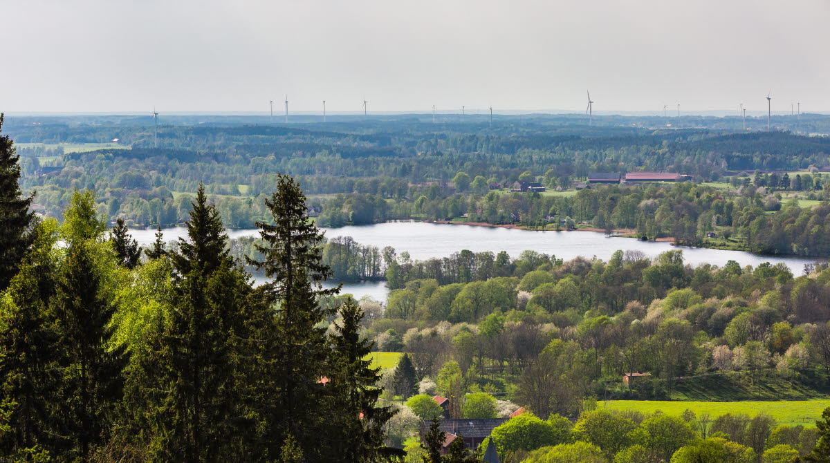 Utsikt över Valle med gröna ängar, körsbärsträd som är på väg att slå ut, sjöar och långt bort i horisonten syns, vindkraftverk, berget Kinnekulle och Vänern.
