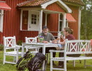 Red cottage. In front of the cottage sits a couple having coffee in white garden furniture.