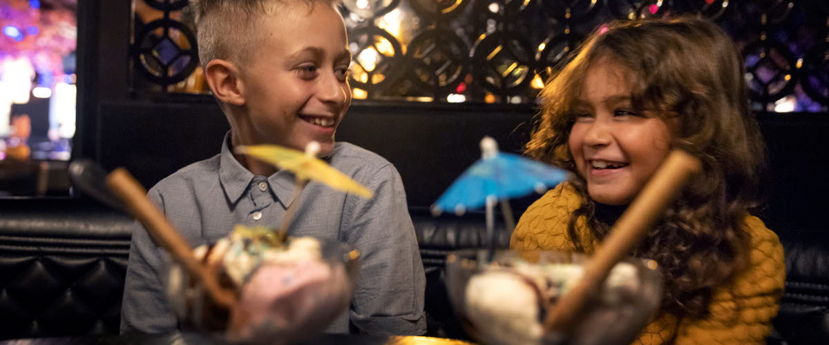 Två barn, en kille och en tjej, sitter på restaurang med två festliga efterrätter framför sig. 