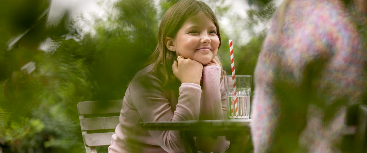 En tjej sitter vid ett bord och fikar i en mysigt lummig miljö bland trädgårdsbuskar och röda husfasader. 
