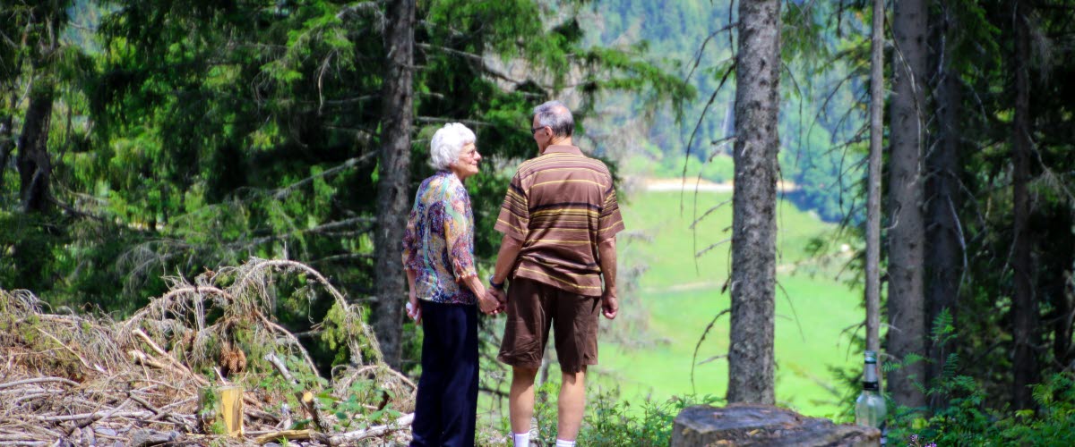 Två äldre personer står i en skogsdunge, håller varandra i händerna och tittar på varandra. 