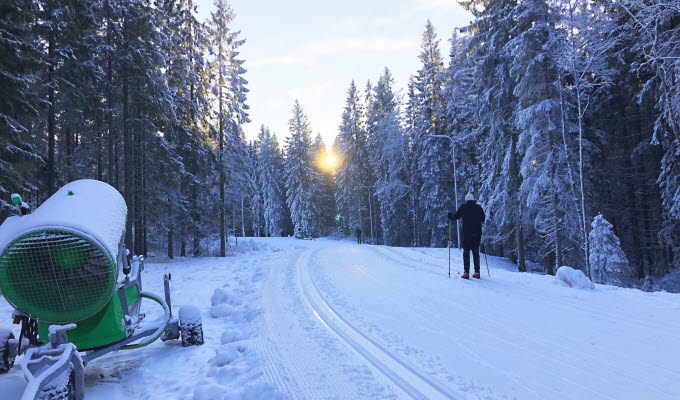 Ensam skidåkare i längdskidspåren på Billingen. Solen glittrar genom de snötäckta träden och snökanonen står redo på sidan av spåren.