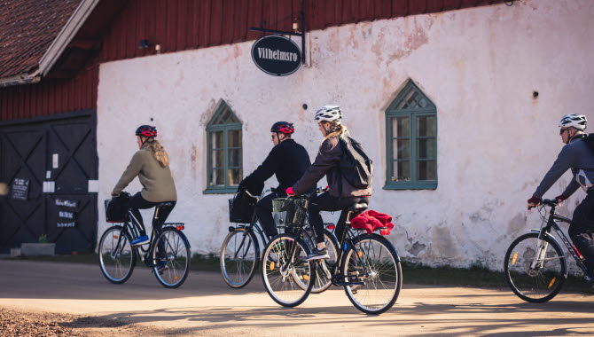Fyra personer som cyklar förbi en ladugårdsbyggnad med en skylt Vilhelmsro på väggen.
