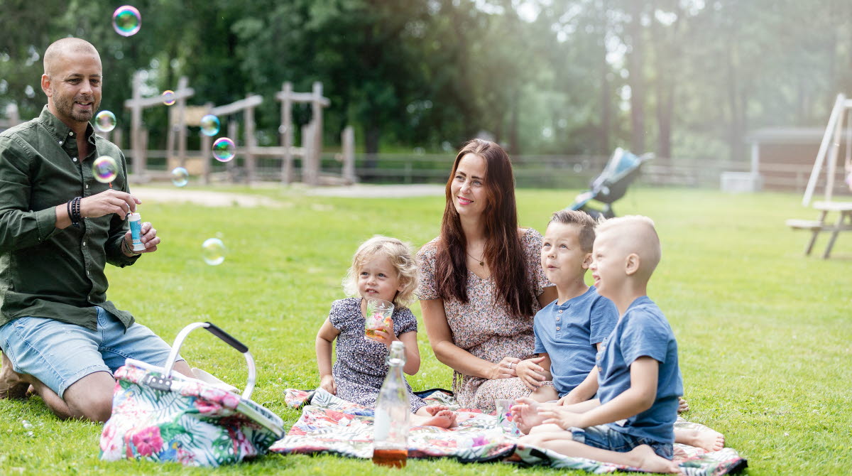 Pappa som blåser såpbubblor, tre barn och mamman sitter på en filt på gräset och ser på och fika.