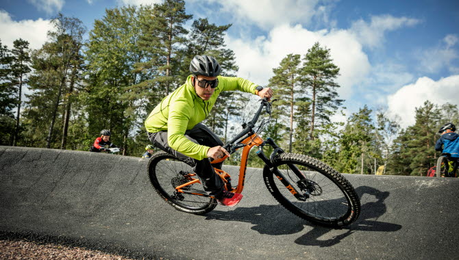 En kille i neongul jacka, solglasögon och svart cykelhjälm cyklar på sin orangea cykel på en asfaltsbana, en pumptrackbana. I bakgrunden ser man Billingens skogar.