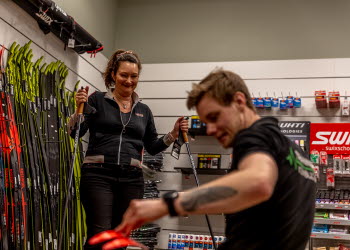 Kvinna står med stavar i en sportbutik. En manlig expedit sitter och plockar fram något till henne. I bakgrunden är en hel vägg med längdskidor.