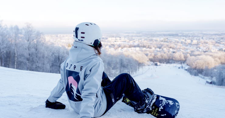 Person with helmet and snowboard sits in Billingebacken and looks out over the hill and the town of Skövde