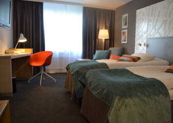 Två sängar med turkosa överkast står intill varandra inne på ett hotellrum. Framför sängar står även ett skrivbord med en orange stol.