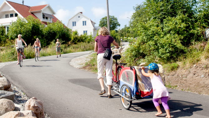 En mamma och ett barn kämpar sig upp för en backe vid Ekenäs med en cykel och en cykelkärra. Mamman leder cykeln och barnet puttar på kärran.