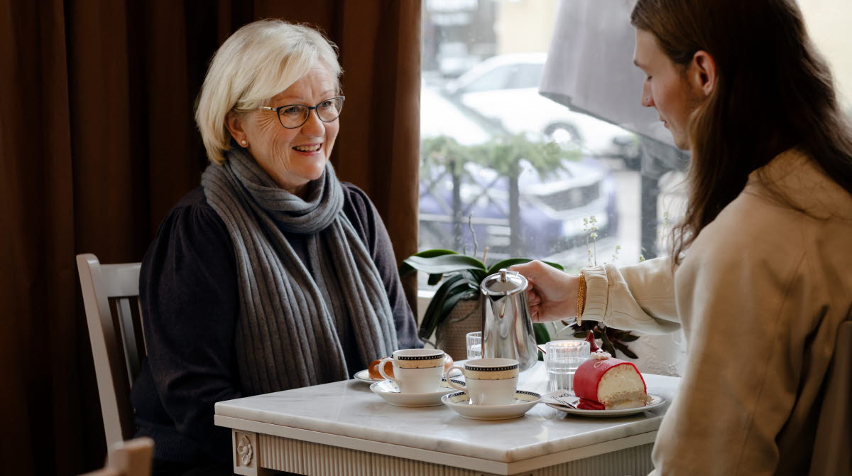 En äldre dam sitter och fikar vid ett bord med en yngre person i vit tröja och långt hår som häller upp varm dryck från en kanna.