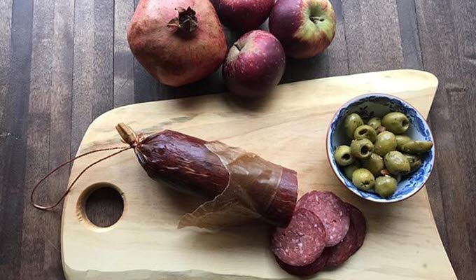 En bild på ett bord med en skärbräda där det ligger en rejäl korv som någon skurit upp några skivor av. På skärbrädan står också en liten porslinsskål med oliver. Jämte skärbrädan ligger röda äpplen och ett granatäpple.