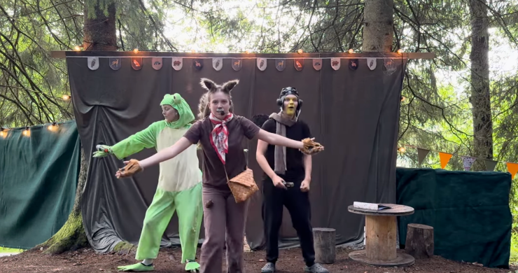 Tre karaktärer – en groda, en varg och en fågel – rör sig energiskt mot kameran på en scen i skogen, som en del av ett färgstarkt utomhusframträdande.