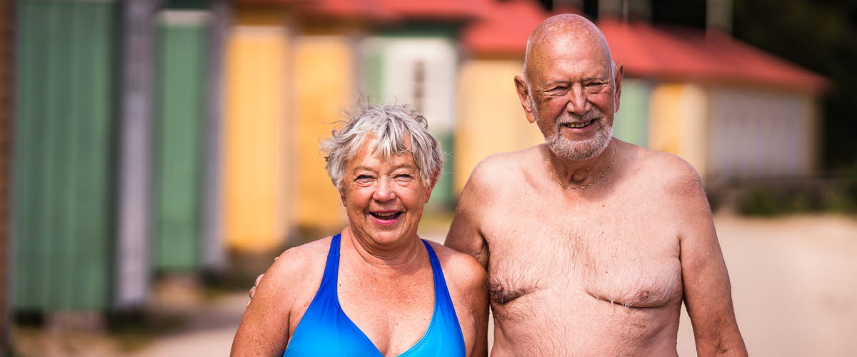 Äldre par vid Guldkroksbadets sandstrand, framför badhytter.