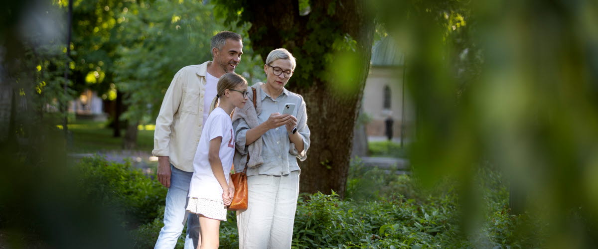 A man, a woman, and a girl are standing in a park, looking down at a phone.