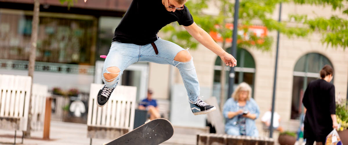 En kille i svart tshirt och ljusa jeans med slitningar på knäna gör ett trick på en skateboard på torget.