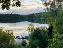 En man står vid kanten av en spegelblank sjö och fiskar i skymningen. 