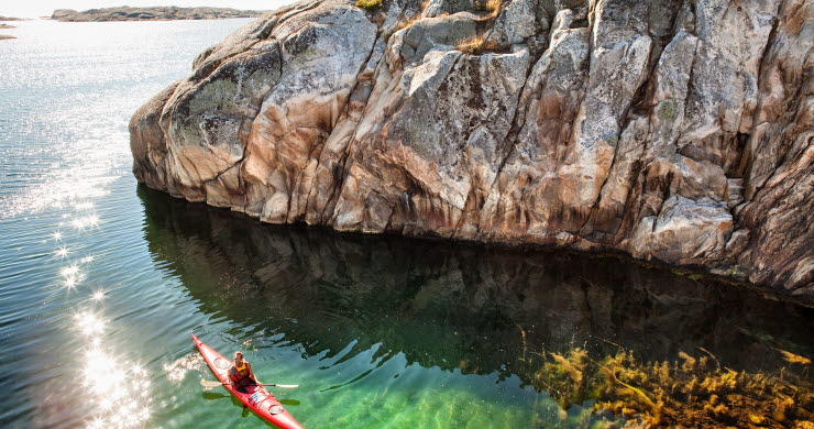 En röd kajak paddlar förbi en klippvägg. Det är en solig dag och vattnet är klart. 