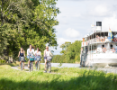 Four women cycle in the green beautiful surroundings along the Göta Canal. Next to them in the canal there is a passenger steamboat.