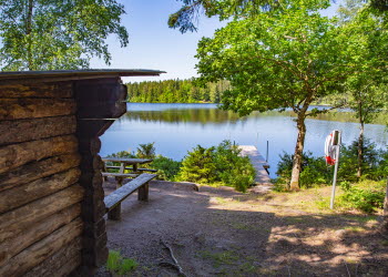Fiskesjön Hållsdammen med ett vindskydd i trä till vänster i bild. Det är lummigt och grönt. 