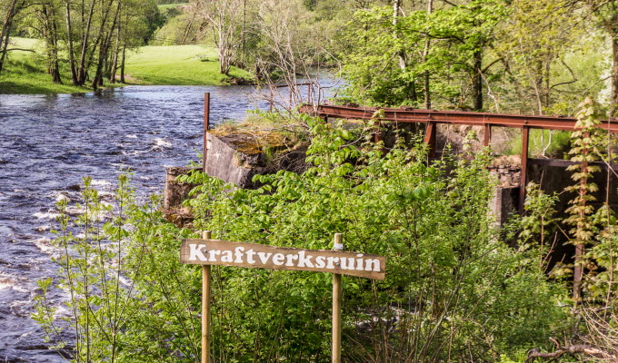 En bild på en strömmande å i ett grönt landskap. Vid ån sitter en träskylt med texten "Kraftverksruin".