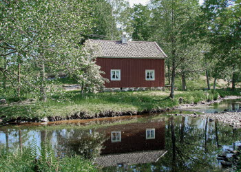 En bild på ett rödmålat hus som står i ett grönt landskap bredvid en spegelblank å.
