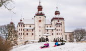 Två barn åker pulka från en backa framför slottet Läckö Slott. Marken är täck av snö. 