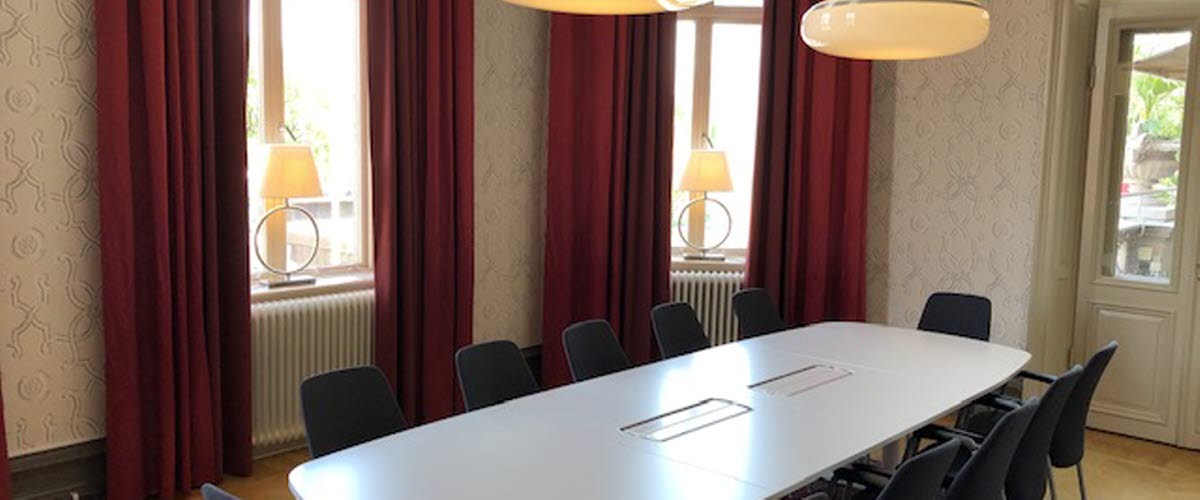 Konferensrum med röda gardiner vid de stora fönstren, vitt bord och grå stolar på hotell Scandic Billingen. 