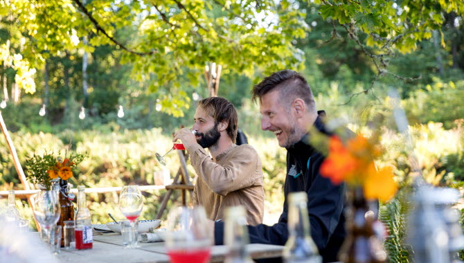 Två män äter och skrattar vid bord ute under ett stort träd.