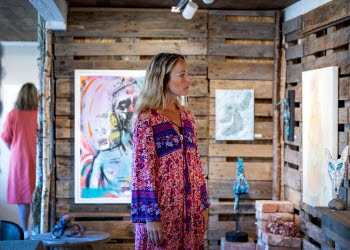 En kvinna i blont långt hår och mönstrad långklänning står och beundrar konst i en konsthall