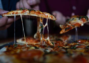 En närbild på händer som plockar varsin pizza slice från en pizza. Det är gott om osttrådar.