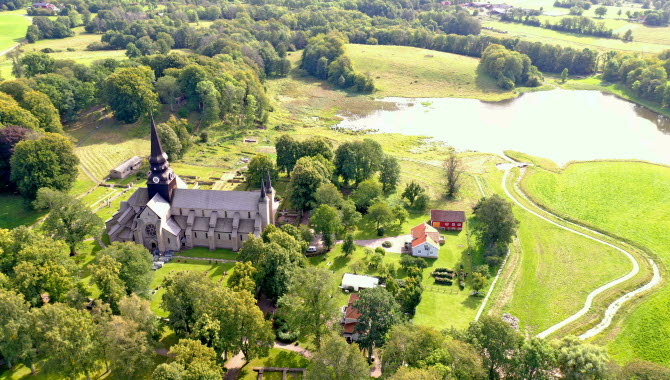 Flygbild över ett grönskande Varnhem. Till vänster i bild syns en klosterkyrka och till höger en spegelblank liten sjö. Runtom ligger små boningshus.