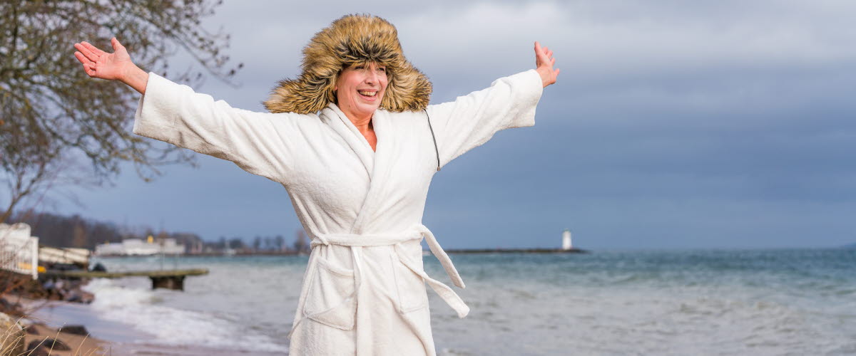 Kvinna står på strand i Hjo mitt i vintern, i morgonrock och pälsmössa. 