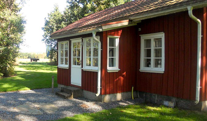 En närbild på ett rödmålat torp med vita spröjsade fönster. Huset är omgivet av grönska.